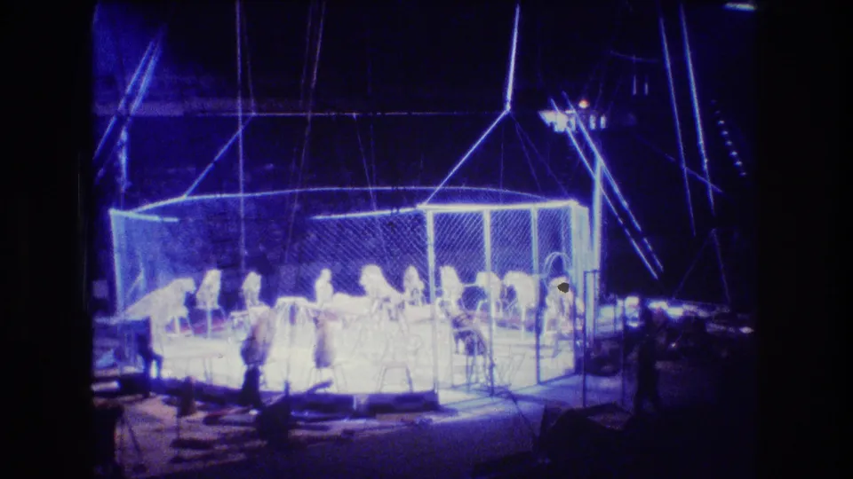 The circus ring remains brightly illuminated in blue, with distant people and likely lions visible inside the cage against a mostly dark background.