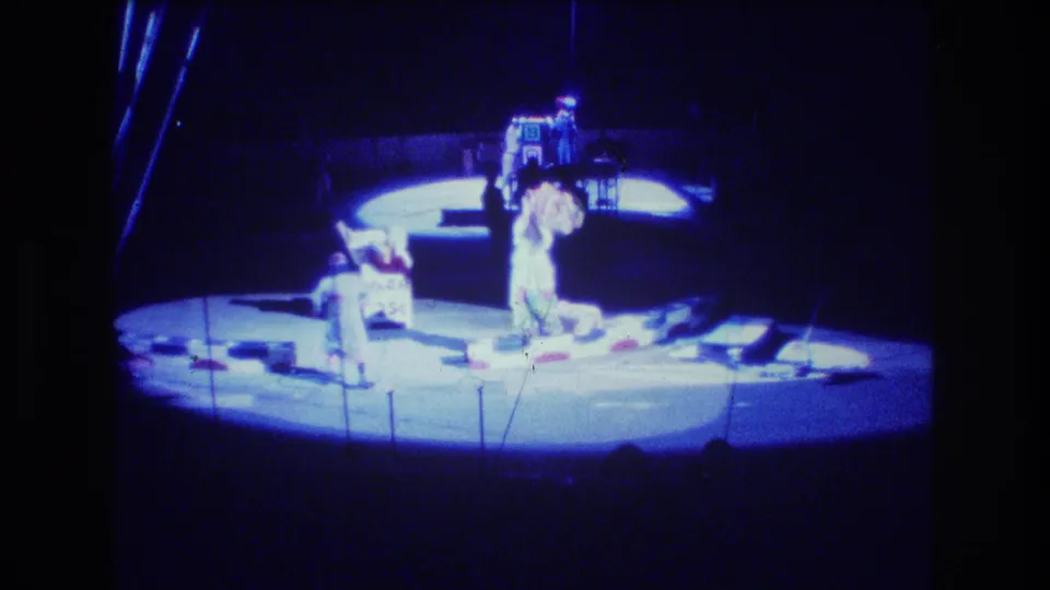 1974 Boston 8mm frame showing a circular circus stage under blue spotlights, with several costumed performers or trainers positioned around props and stands.