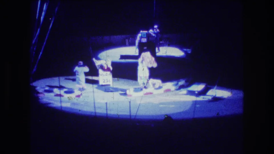 1974 Boston 8mm frame showing performers or trainers on a circular stage with a sign-like prop, scattered floor props, and heavy blue-white exposure from the spotlight.
