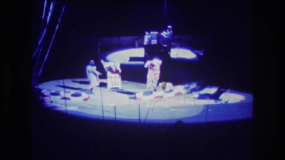1974 Boston 8mm frame showing a distant audience view of a circus-style stage, with costumed performers near props and a sign-like board under blue theatrical lighting.