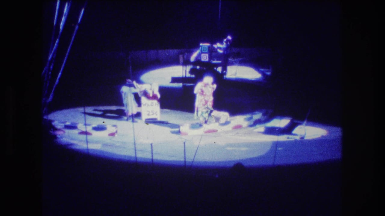 1974 Boston 8mm frame showing costumed circus performers under blue stage lighting, with a small 25 cent sign near the center of the circular stage.
