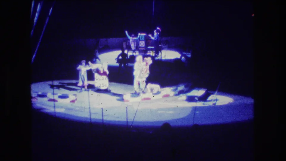 1974 Boston home-movie frame of a circus stage act, with performers spaced around a circular lit area and dark seating or backstage space surrounding them.