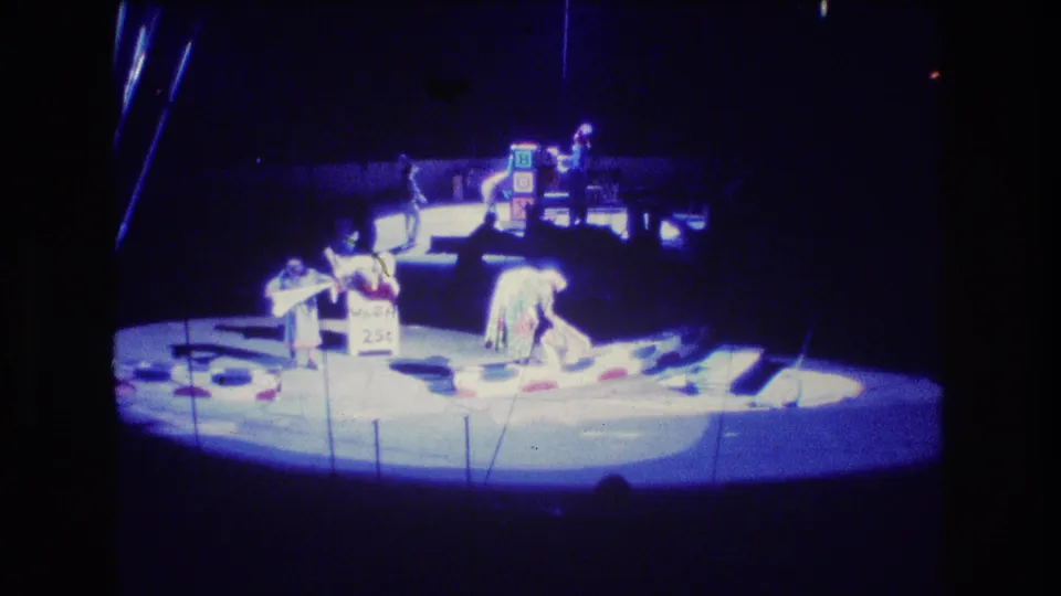 1974 Boston 8mm frame showing performers mid-routine on a circular circus stage, with props on the floor and a 25 cent sign visible near the center.