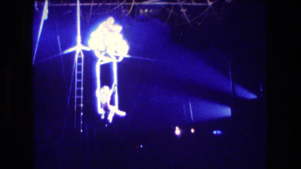 Boston 1974 circus frame with acrobats on an elevated rig, one figure hanging below while bright stage light and blue exposure dominate the dark arena background.