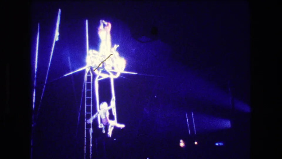Blue-lit 8mm frame from the Boston circus performance showing acrobats on and below an elevated structure, with ladder, ropes, and strong stage glare softening the figures.