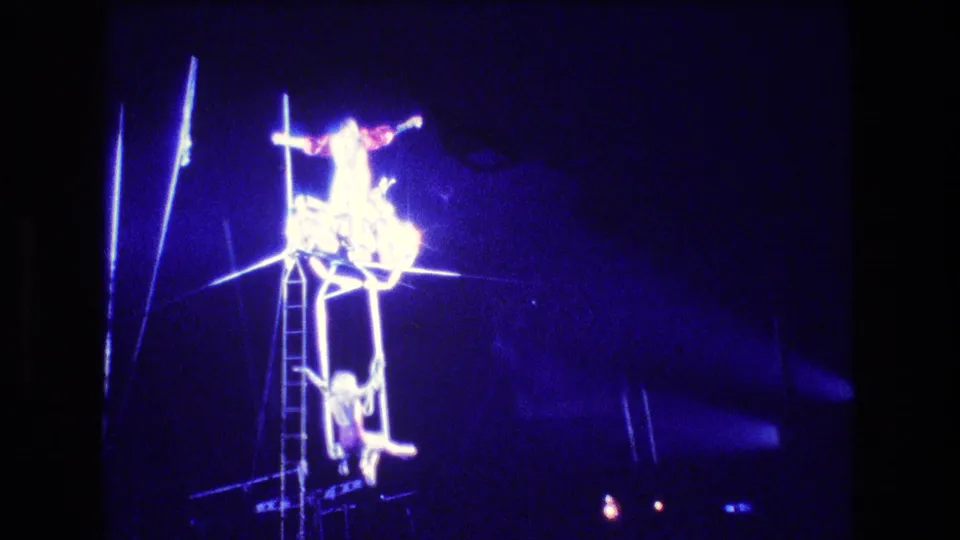 Boston 1974 8mm frame of the aerial act at height, with one acrobat extended on the upper rig, another below, a ladder at center, and blue spotlights cutting through the dark venue.