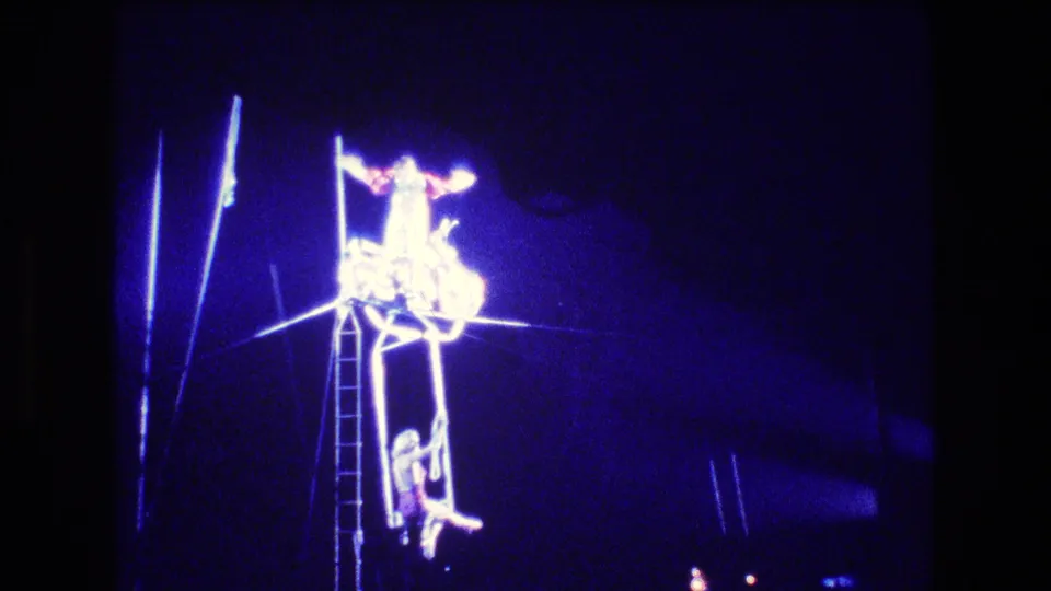 Boston 1974 circus scene with a costumed acrobat positioned high above the ring near a ladder and aerial rigging.