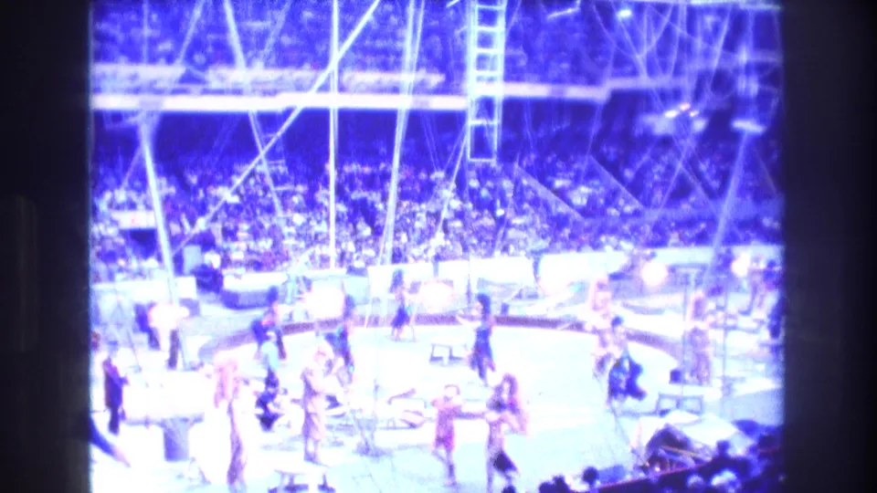 Wide Boston 1974 circus ring view with performers on the floor, overhead rigging, a ladder, and a seated audience under bright blue lighting.