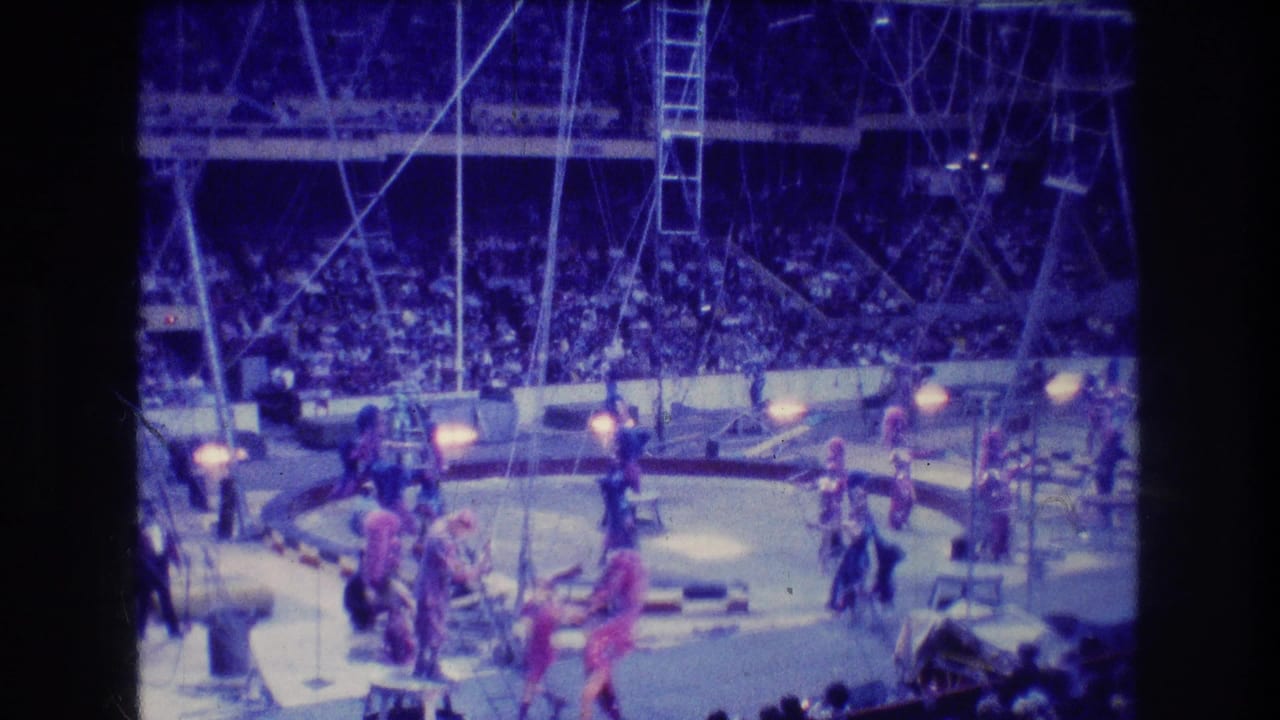 1974 Boston 8mm frame showing costumed circus performers spread across a circular ring, with rigging, lights, and a large seated audience in the background.