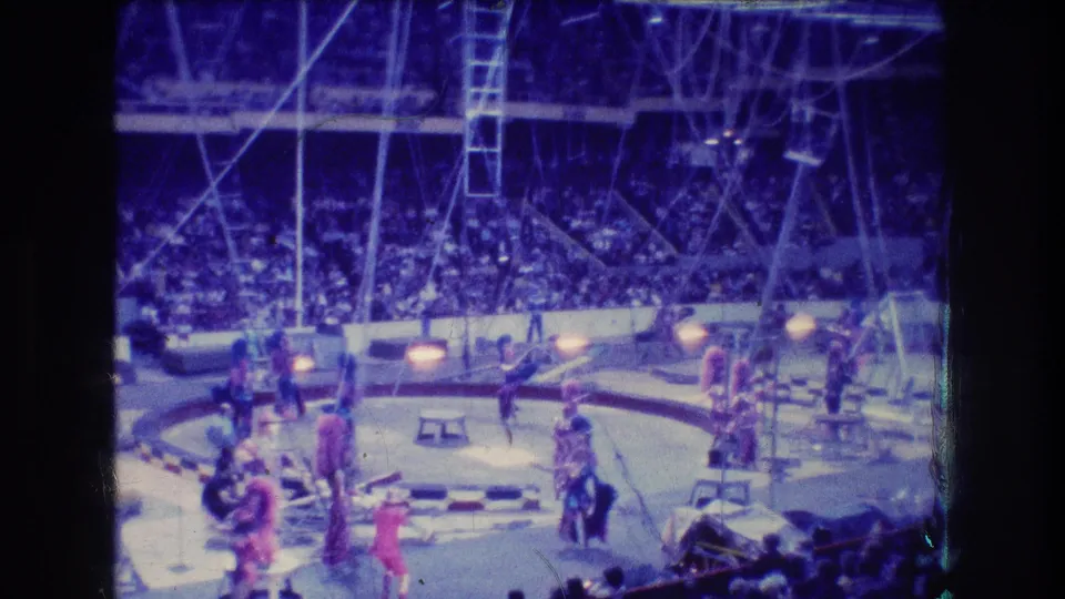 Audience-view 1974 Boston circus frame with dancers or acrobatic performers in bright costumes, props on the ring floor, rigging above, and spectators filling the stands.