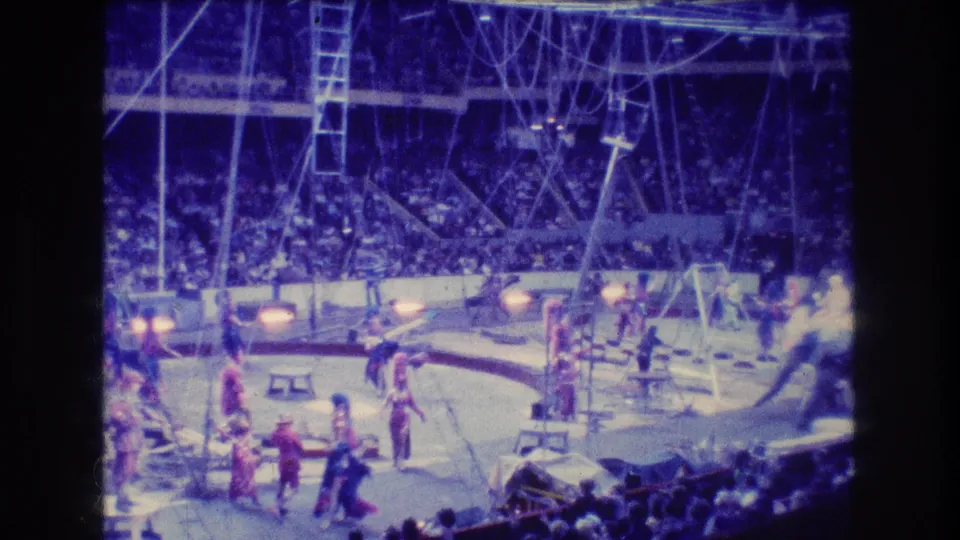 1974 Boston home-movie frame showing a wide view of the circus ring with multiple performers, visible rigging, a ladder structure, props, and a crowded arena behind them.