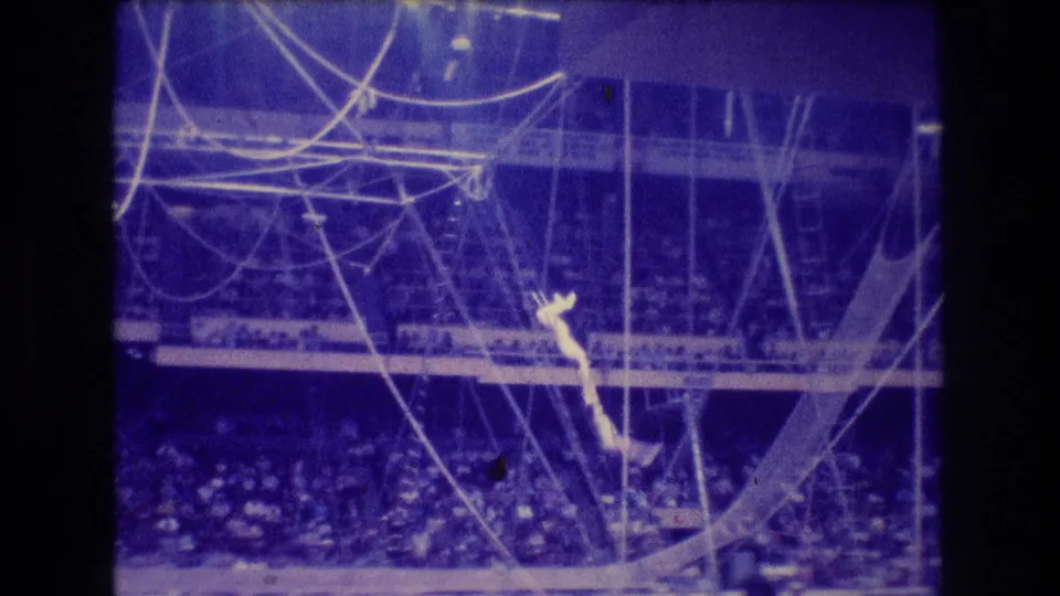 1974 Boston 8mm frame showing a distant trapeze performer hanging downward amid ropes and cables, with the audience visible below in soft blue grain.