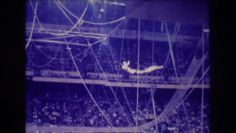 1974 Boston circus frame showing two distant aerial performers extended near trapeze ropes above a crowded arena, with strong blue color shift and visible film grain.