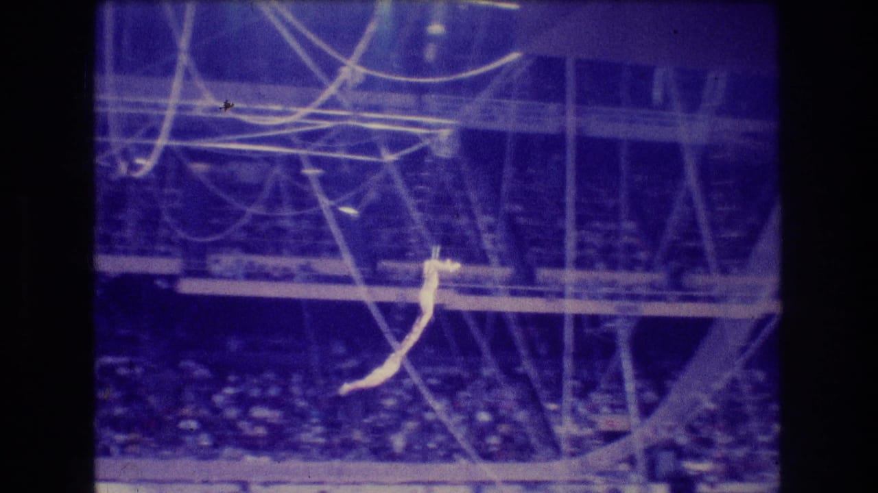 1974 Boston 8mm frame showing a light-clad trapeze artist suspended under circus rigging above a darkened arena audience in blue-purple light.
