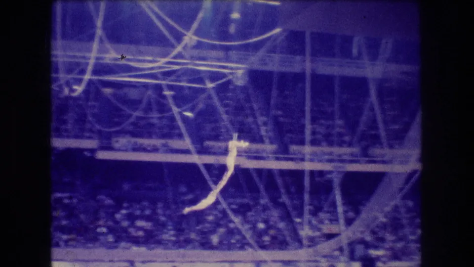 1974 Boston 8mm frame showing a light-clad trapeze artist suspended under circus rigging above a darkened arena audience in blue-purple light.