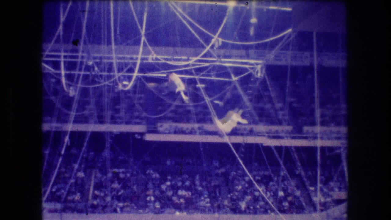 1974 Boston 8mm frame with two distant trapeze artists amid overhead ropes and bars, with the audience visible below in blue stage lighting.