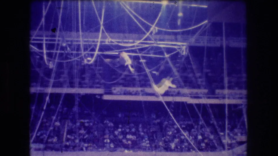 1974 Boston 8mm frame with two distant trapeze artists amid overhead ropes and bars, with the audience visible below in blue stage lighting.