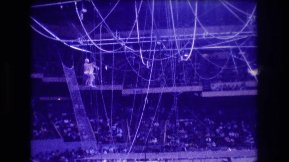 1974 Boston 8mm frame showing a distant trapeze performer near suspended ropes and netting above a seated arena crowd under blue-purple circus lighting.