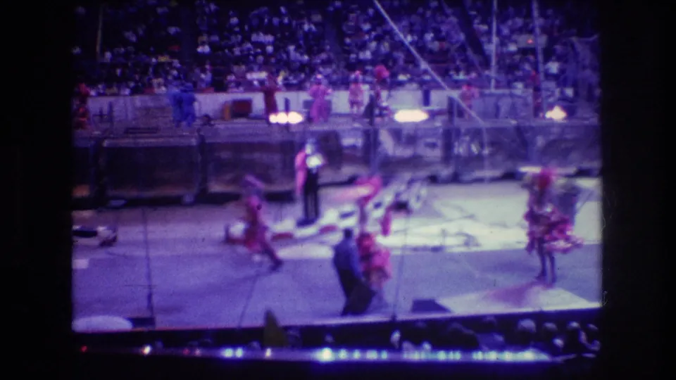 Costumed circus workers cross the lit arena floor in Boston while audience members sit in the background and ring equipment remains visible.