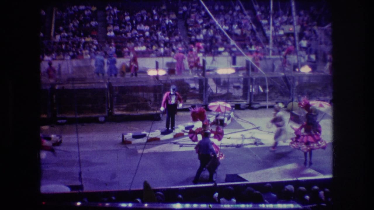 Circus performers in red costumes move through an arena ring as a suited ringmaster stands nearby.
