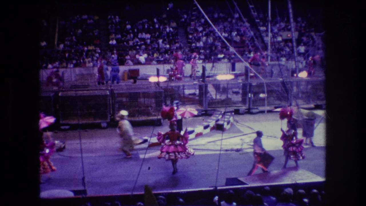 Circus performers with umbrellas cross the ring while spectators fill the stands behind them.