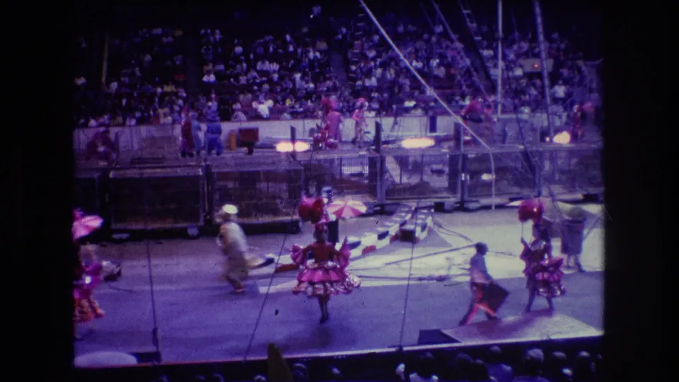 1974 Boston 8mm home-movie frame of a circus act with red-costumed performers carrying pink umbrellas under arena lighting, viewed from the audience seating.
