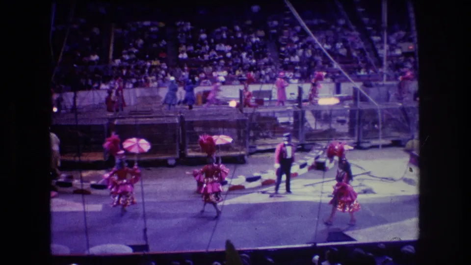 1974 Boston 8mm home-movie frame showing the ringmaster near center ring while several red-costumed performers with umbrellas continue a circus routine before the crowd.