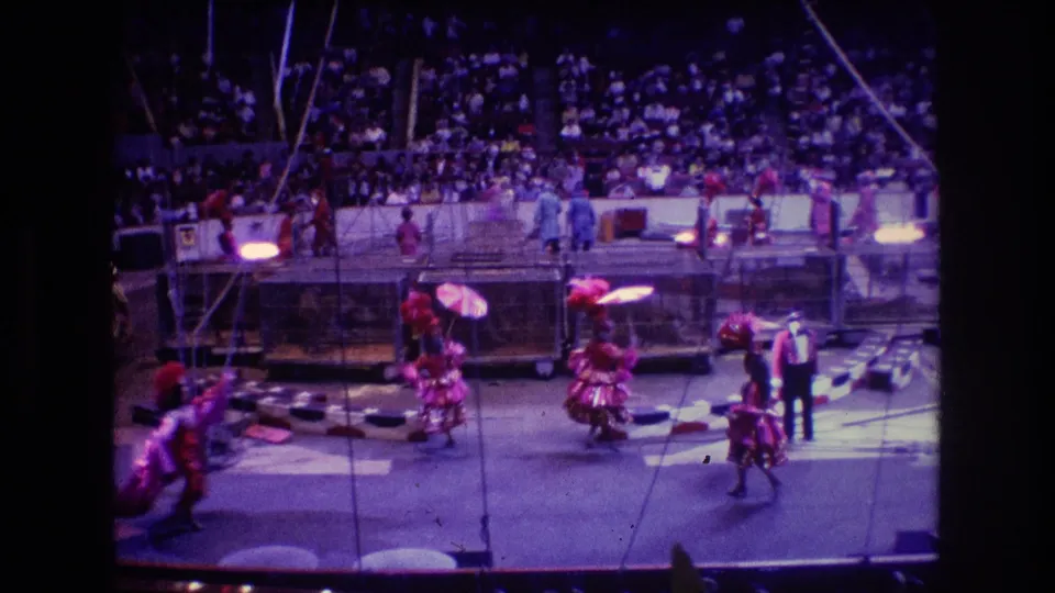 1974 Boston 8mm home-movie frame showing a wider audience-shot view of umbrella performers in red costumes moving through the circus ring with the ringmaster nearby.
