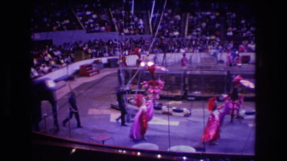 Boston 1974 circus footage shows dancers or performers with parasols near rigging and arena barriers, filmed from a distant audience viewpoint.