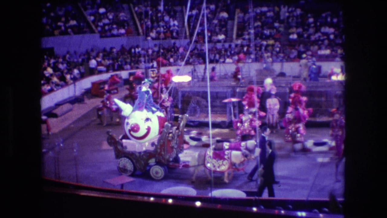 Clown-faced cart pulled by horses in a circus ring with performers and audience seating behind it.