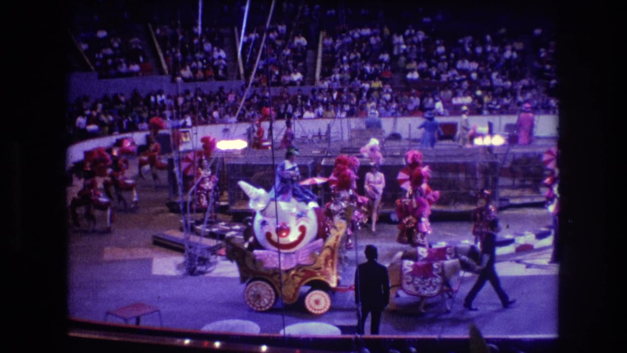 Clown cart and horses move through the ring while performers stand near circus equipment and spectators fill the seats.