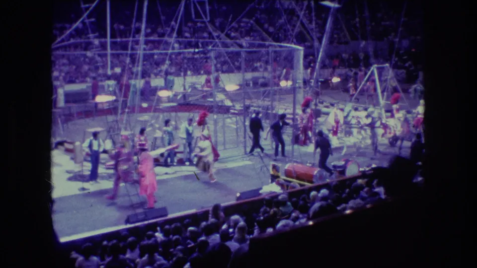 1974 Boston home-movie frame showing a broader circus arena view with performers, apparatus, audience seating, and elephants near the side of the ring.