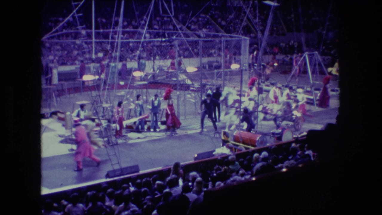 Wide 1974 Boston home-movie view of an indoor circus performance with costumed performers, metal rigging, props, and audience seating under purple-blue stage light.