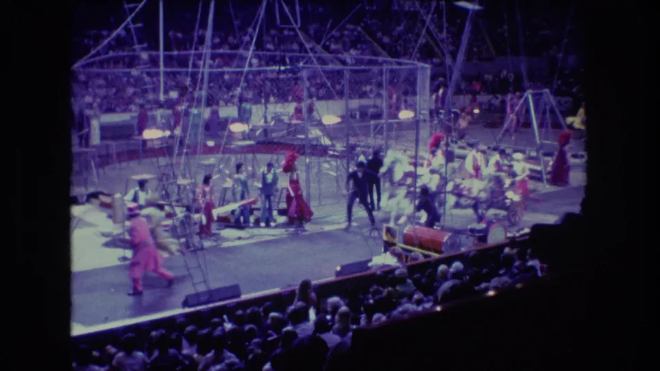 Wide 1974 Boston home-movie view of an indoor circus performance with costumed performers, metal rigging, props, and audience seating under purple-blue stage light.