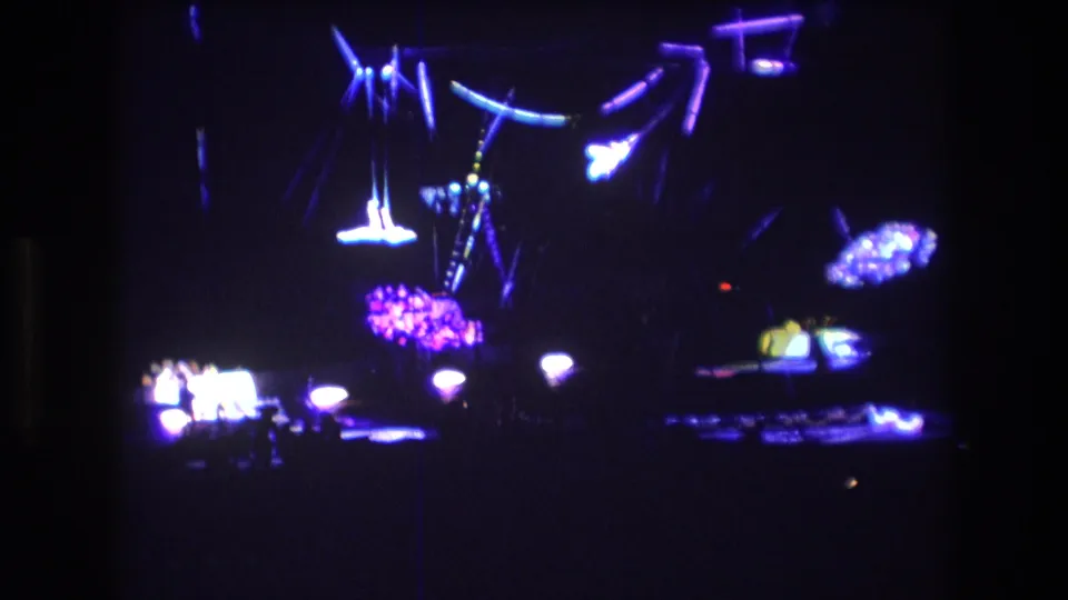 Boston 1974 home-movie frame with suspended circus rigging, intense purple-blue lighting, and small indistinct performers or stage objects below.