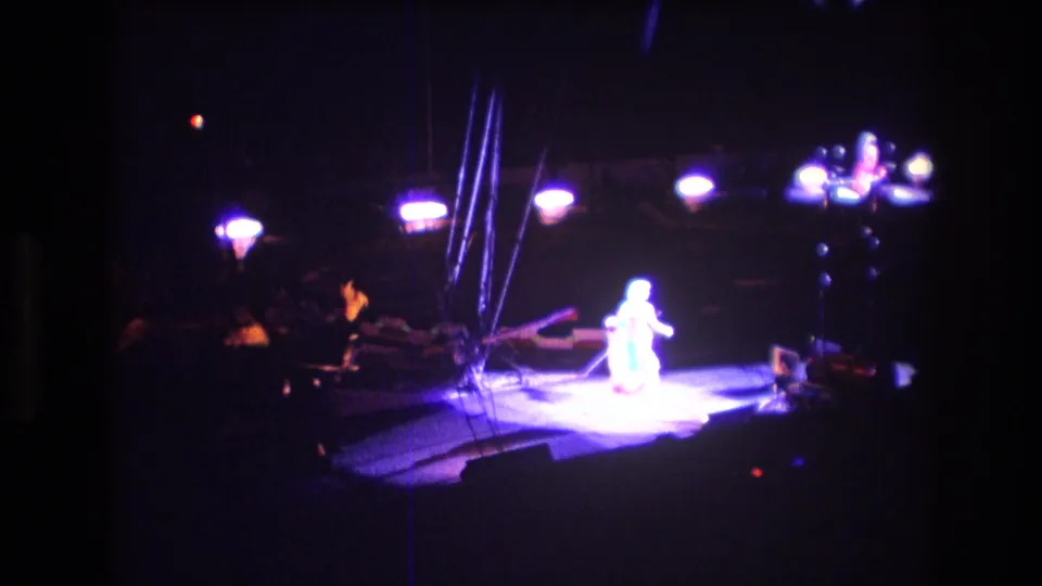 Boston 1974 8mm frame showing a performer in a strong spotlight on a circus stage, with aerial rigging and dim surrounding figures nearby.