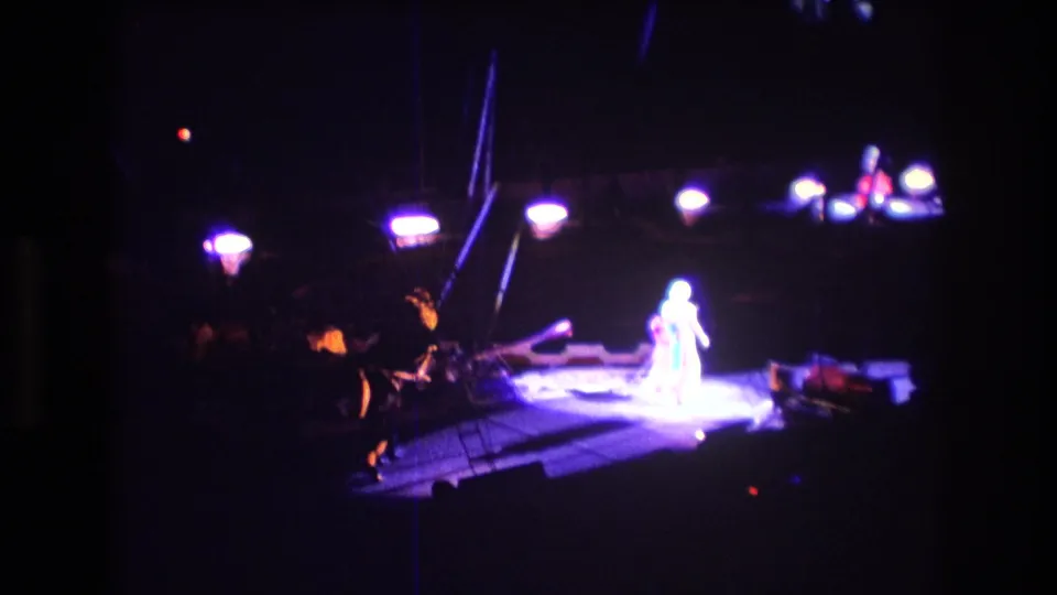 Boston 1974 8mm home-movie frame showing a bright white-costumed circus performer isolated by spotlights on a raised center-ring platform, with surrounding equipment fading into darkness.