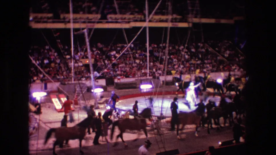 Boston 1974 8mm home-movie frame showing a wide view of the circus ring with horses and handlers in motion, overhead rigging, bright floor lights, and a densely seated audience behind them.