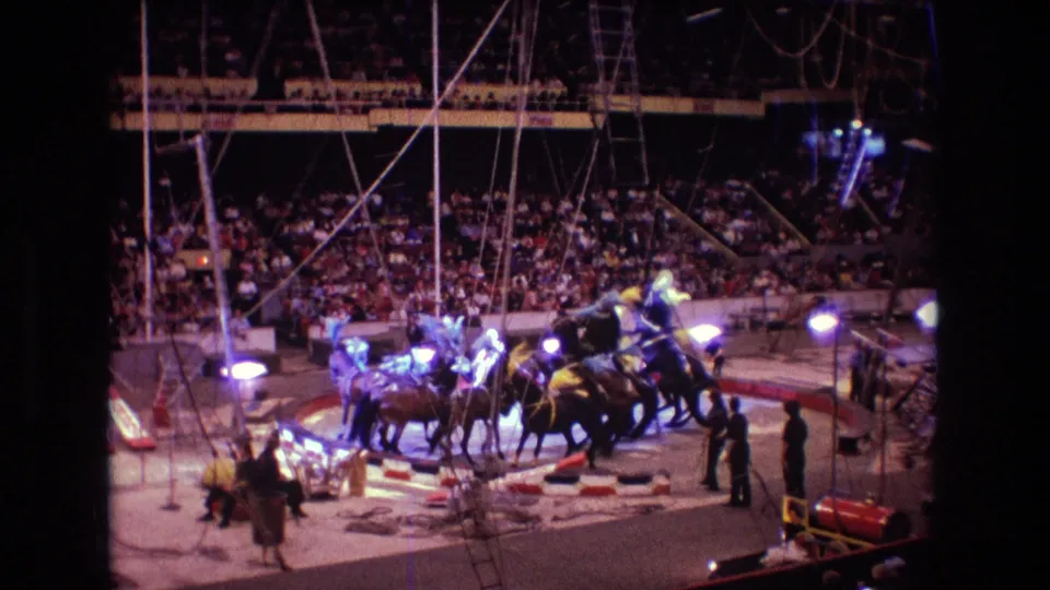 1974 Boston 8mm still of riders and horses moving across a circus ring, with spotlights, props, attendants, and a large seated crowd visible behind them.