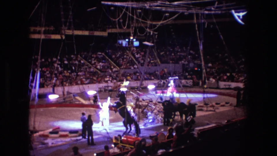 Boston 1974 home-movie still showing a circus horse rearing in the arena as a performer and attendants stand near the ring under overhead rigging.