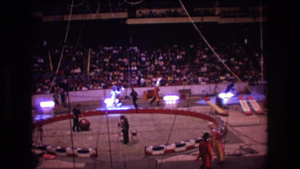 1974 Boston 8mm frame showing a circus ring viewed from the stands, with performers under spotlights, a large red ball, rigging lines, and a seated audience behind the barrier.
