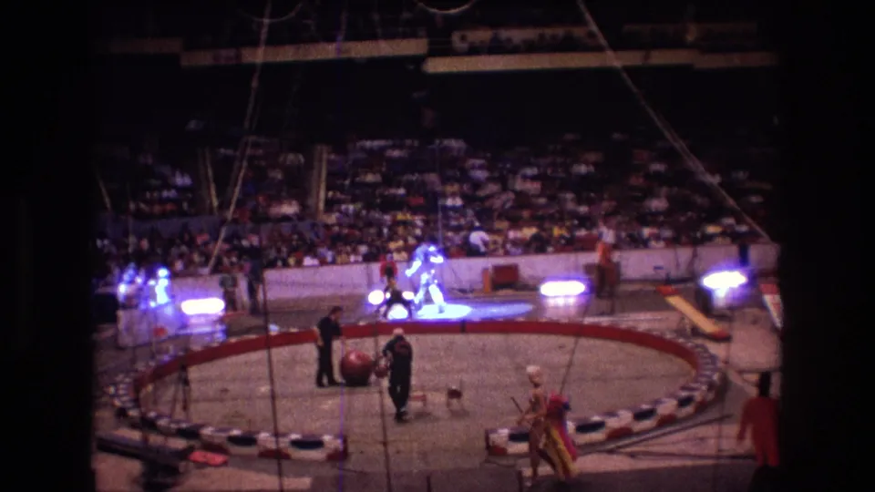 1974 Boston 8mm frame showing a spotlit circus act inside the ring, with clowns or costumed performers, arena seating, and visible rigging above the performance area.