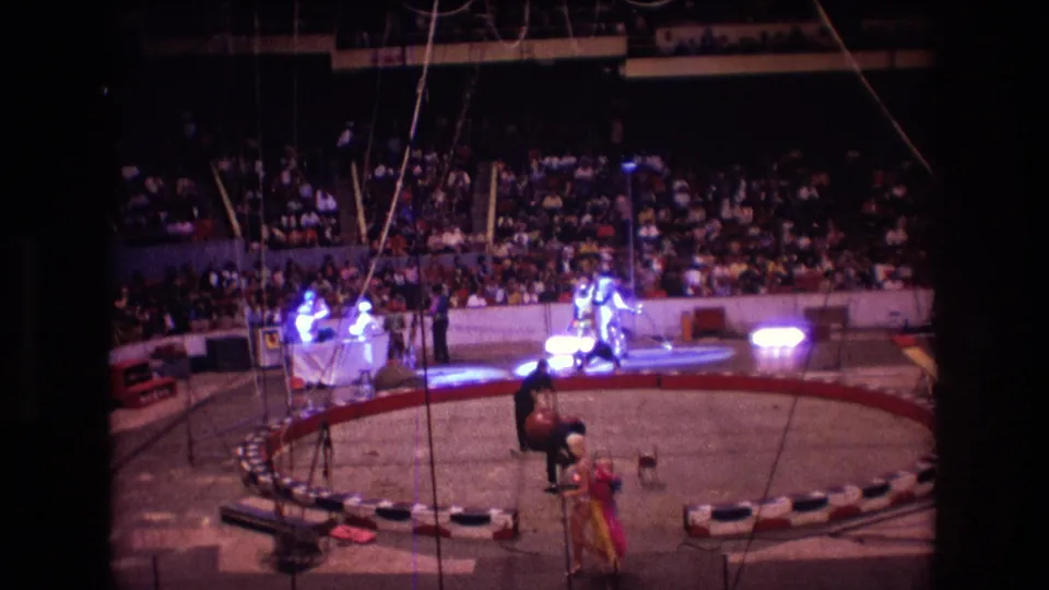 1974 Boston 8mm frame showing circus performers near a large red ball, with the circular ring, bright stage lights, red-and-white edging, and a crowded arena visible in the background.