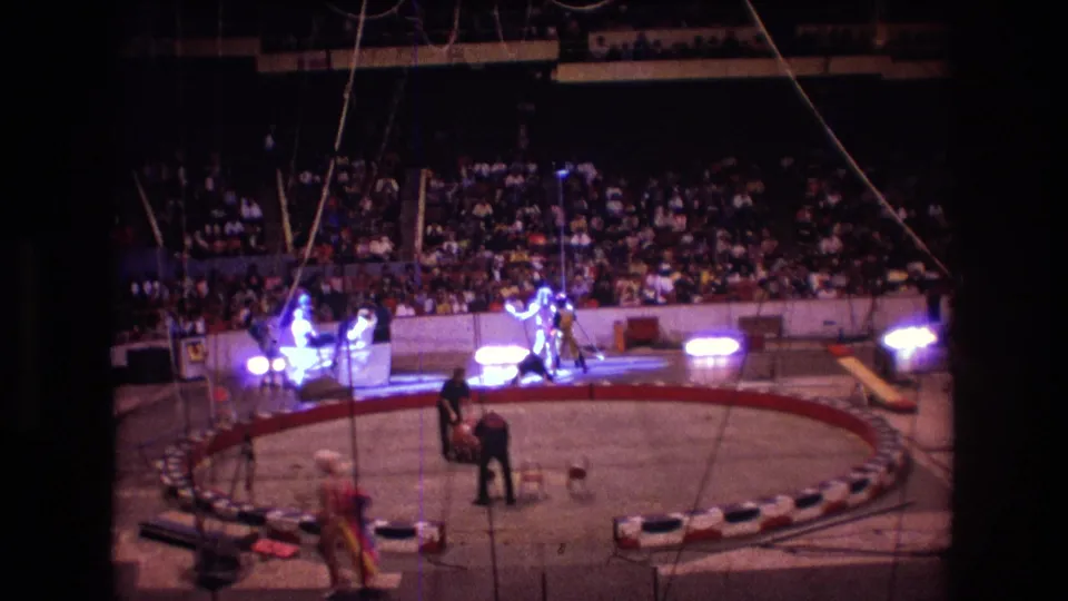1974 Boston 8mm frame showing performers moving through a lit circus ring, with overhead rigging, a large red ball, red-and-white ring barrier, and spectators filling the arena seats.