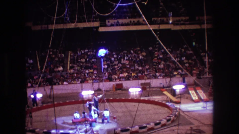 Boston 1974 8mm frame showing assistants gathered beneath a tall circus apparatus as a spotlighted performer appears elevated above the ring in front of the seated audience.