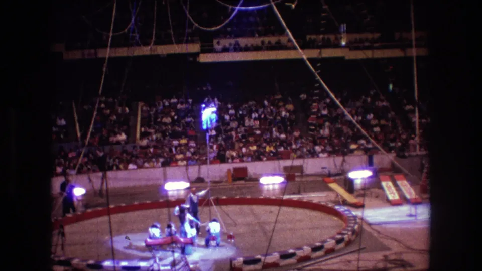 Boston 1974 frame showing the spotlighted circus performer high above the ring, with assistants at floor level and a large audience visible in the dim arena background.