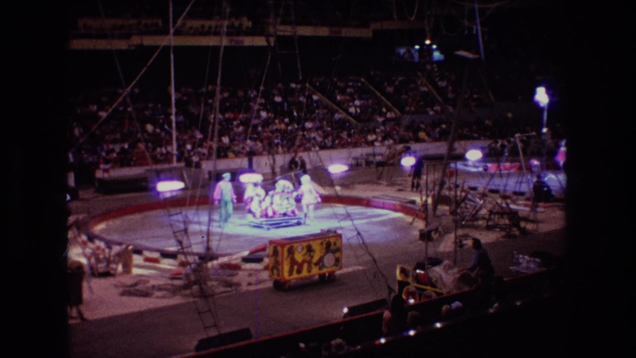 Circus ring with spotlit performers, audience seating, and a decorated wagon at the edge of the arena.