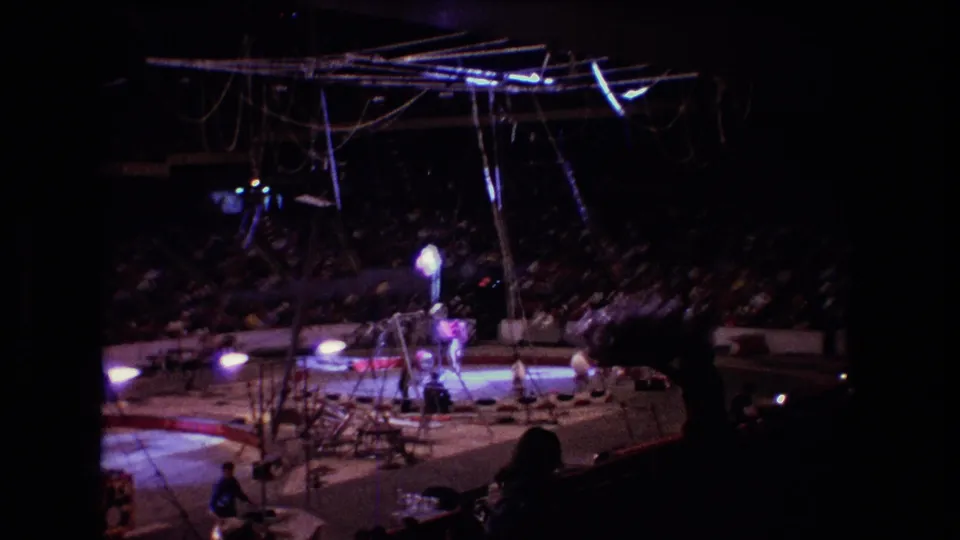 1974 Boston circus footage frame from the stands, with aerial rigging above the ring, performers under bright lights, and a crowded audience in the background.