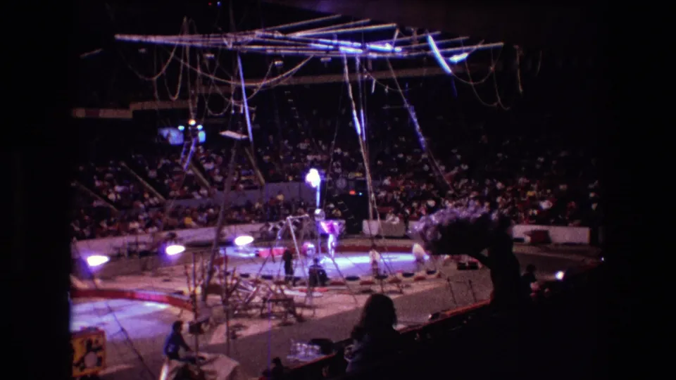 1974 Boston home-movie frame showing circus apparatus suspended above the ring, audience members in the stands, and a silhouetted spectator near the camera position.