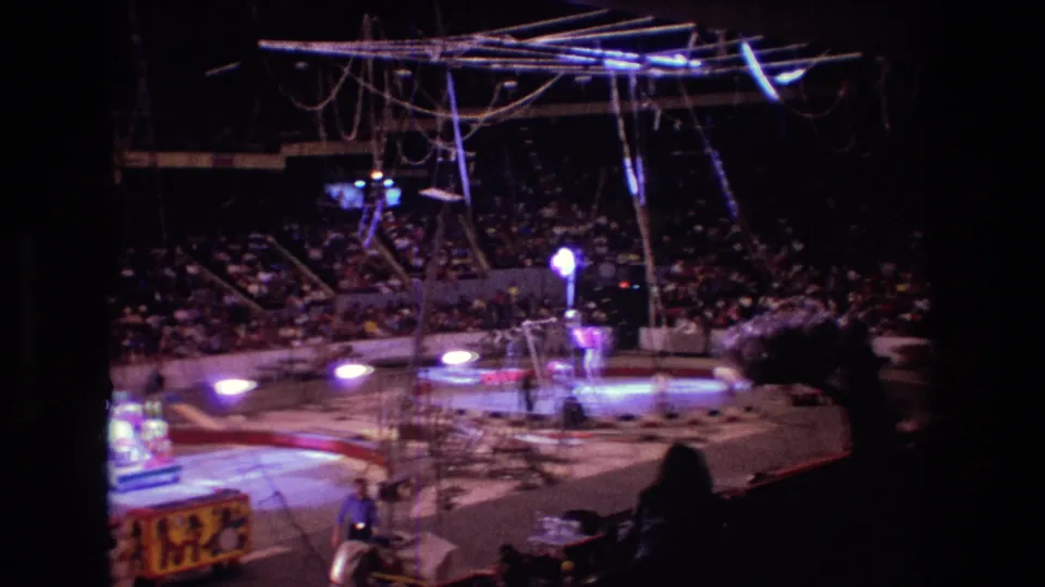 1974 Boston 8mm frame capturing the circus ring from a seated audience viewpoint, with aerial equipment overhead, spectators in the stands, and a decorated wagon partly visible below.